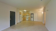 This single-floor unit offers a practical and functional open-concept layout, seamlessly connecting the kitchen and living areas into one cohesive space. The kitchen is well-appointed with light wood cabinetry, a white electric range, a refrigerator, and ample counter space arranged in an L-shaped configuration. An overhead fluorescent light fixture illuminates the kitchen workspace, while a range hood above the stove provides ventilation. A standout feature of this unit is the in-unit stacked washer and dryer, conveniently tucked between the kitchen and an interior door, adding significant everyday convenience. The living area is generously sized, finished with wall-to-wall carpeting that creates a warm and comfortable atmosphere. The light-toned walls throughout contribute to a bright and airy feel. Multiple interior doors provide access to additional rooms or storage spaces, enhancing the overall functionality of the layout. The unit is finished with standard ceiling heights and tile flooring in the kitchen transition zone, offering a clean and easy-to-maintain surface. Overall, this space delivers a straightforward and efficient floor plan well-suited for comfortable day-to-day living.