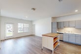 This single-floor residence offers a thoughtfully arranged layout comprising two bedrooms, two bathrooms, a combined kitchen and living area, two closets, and a dedicated laundry room. The open-concept kitchen and living space serves as the heart of the unit, finished with light wood-look luxury vinyl plank flooring that extends throughout the main areas. The kitchen is equipped with stainless steel appliances — including a refrigerator, range, microwave, and dishwasher — complemented by grey cabinetry, light stone-look countertops, and a white subway tile backsplash. A mobile kitchen island with a butcher block top provides additional prep and storage space. The living area benefits from natural light through multiple windows fitted with horizontal blinds. Both bedrooms feature plush carpet flooring and ceiling fans, with each room accessing a dedicated closet. The primary bedroom includes a walk-in closet. The bathrooms are clean and functional, with one featuring a walk-in shower. A separate laundry room houses a full-size top-load washer and dryer. The unit maintains a bright, neutral palette throughout, creating a clean and cohesive living environment.