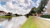This single-story residence is situated within a well-maintained Florida community distinguished by its tranquil waterfront setting. The exterior environment is a standout feature, with a calm canal running alongside the property, offering unobstructed water views framed by lush, mature landscaping. Towering palm trees, broad shade trees, and neatly trimmed hedges contribute to the tropical character of the surroundings, while an expansive green lawn borders the water's edge.

The building itself displays a warm, earthy stucco exterior typical of Florida residential architecture, with arched garage bays providing covered parking directly accessible from a paved lot. A pedestrian walkway runs along the canal, offering a pleasant outdoor corridor beneath a canopy of trees. The community's layout emphasizes open-air living and natural scenery, with wide skies and cloud-filled horizons adding to the airy, sun-drenched atmosphere.

The setting reflects a community designed for comfortable Florida living, combining practical amenities such as attached garages with scenic outdoor spaces. The waterfront canal serves as the defining natural feature, providing a peaceful backdrop and a strong connection to the lush, subtropical landscape characteristic of the region.