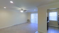 This single-floor residence features a well-appointed living area finished with neutral-toned carpeting that extends throughout the main space, creating a clean and cohesive look. Crown molding trims the perimeter of the ceiling, adding a classic architectural detail that elevates the overall aesthetic. A ceiling fan with a built-in light fixture is centrally mounted, providing both air circulation and ambient lighting. Recessed lighting complements the ceiling fan, ensuring the space is well-lit. A sliding glass door, dressed with vertical blinds, serves as a prominent feature along one wall, allowing natural light to filter into the room while offering access to the exterior. Baseboard heating runs along the lower walls, supplemented by a wall-mounted HVAC unit positioned near the sliding door, providing year-round climate control. The walls are painted in a soft, warm neutral tone, contributing to a bright and inviting atmosphere. Tile flooring is visible at the entryway, transitioning into the carpeted living area and providing a practical, durable surface near the entrance.