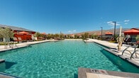 Located in the Rocking K Ranch community in Tucson, Arizona, this single-level outdoor amenity complex offers a resort-caliber experience set against a dramatic Sonoran Desert mountain backdrop. At the heart of the space is a large, sparkling resort-style swimming pool with crystal-clear turquoise water, surrounded by an expansive pool deck furnished with lounge chairs and vibrant orange shade umbrellas, providing ample space for relaxation under the Arizona sun. Adjacent to the pool, a manicured artificial turf lawn serves as a versatile recreation area, complete with cornhole boards for casual outdoor gaming and gatherings. Wide concrete pathways connect the various zones of the complex, creating a cohesive and accessible layout throughout. Thoughtfully placed desert-adapted trees and low-water landscaping add a touch of natural greenery while complementing the arid surroundings. Architectural structures with bold red and neutral gray exteriors frame the space, lending a modern ranch-inspired aesthetic consistent with the community's identity. Perimeter fencing with open sightlines preserves the breathtaking mountain views, while tall light poles ensure the space remains functional into the evening hours. The overall ambiance is one of active relaxation, community engagement, and seamless indoor-outdoor living in the heart of the Sonoran Desert.