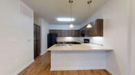 Located at 10765 Highway 78 E in Summerville, South Carolina, this single-story unit offers a well-designed, functional layout with consistent finishes throughout. The open-concept living and kitchen area is a central highlight, featuring warm wood-look flooring that flows seamlessly from the living space into the kitchen. The kitchen is equipped with rich, dark wood cabinetry extending to the upper walls, providing generous storage. Light granite-style countertops offer a pleasing contrast, and a peninsula with a double stainless steel sink and pendant lighting above serves as both a workspace and casual gathering point. Black appliances, including a refrigerator and range, complete the kitchen's cohesive look. A chandelier-style light fixture illuminates the open living area, enhancing the spacious, airy feel of the combined space. The bedrooms are carpeted for comfort, each featuring ceiling fans with light fixtures and windows that invite natural light. One bedroom includes a double-door closet for ample storage. The bathrooms maintain the same design language, with dark wood vanities topped with granite-style counters, full tub-shower combinations, and wood-look flooring. Natural light enters through windows, keeping the bathrooms bright and welcoming.