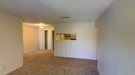 This single-story space in Midland, Texas, offers a straightforward, functional layout designed for comfortable everyday living. The living area is finished with wall-to-wall carpet in a neutral tone, contributing to a warm and understated atmosphere. A textured popcorn ceiling runs throughout, a characteristic feature of the space's construction style.

A notable architectural detail is the pass-through window connecting the living area to the kitchen, framed with a wooden ledge. This opening allows for a sense of visual connectivity between the two spaces while maintaining a defined separation. The kitchen, visible through the pass-through, features wood-toned cabinetry and a range, providing essential cooking amenities.

Toward the rear of the living area, a hallway leads to additional rooms, with a closet visible along the corridor, offering built-in storage. Overhead lighting in the hallway provides adequate illumination throughout. The walls are finished in a neutral off-white tone, keeping the interior feeling clean and bright. Overall, the space presents a practical, no-frills layout well-suited for a variety of living arrangements.