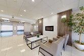 The lobby at 1066 King St W in Kingston, Ontario serves as an elegant and functional gateway to the building. The space is finished with large-format, high-gloss cream tile flooring that reflects the recessed pot lighting overhead, creating a bright and polished atmosphere throughout. A prominent seating lounge anchors the main area, furnished with upholstered armchairs and a sleek white coffee table arranged around a wall-mounted linear fireplace set into a warm wood-paneled feature wall — adding a refined, contemporary touch. Tall indoor plants introduce a natural element and complement the neutral colour palette of beige, grey, and white tones. The entrance features floor-to-ceiling glass doors that draw in natural light from the exterior. Along one wall, a built-in Canada Post mailbox panel and parcel locker station provide convenient mail and package access for residents. Two stainless steel elevators are prominently positioned toward the rear of the lobby, offering efficient vertical access throughout the building. A connecting hallway leads further into the building's interior. The space is illuminated throughout by a combination of recessed pot lights and wall-mounted sconces, ensuring a well-lit and welcoming environment for all who enter.