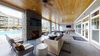 This covered outdoor amenity space is thoughtfully designed to offer a resort-style experience, blending comfort and entertainment in an open-air setting. The architecture is anchored by a dramatic floor-to-ceiling stacked stone fireplace, which serves as the focal point of the lounge area and provides warmth during cooler evenings. A rich, warm-toned wood-planked ceiling spans the entire space, complemented by recessed lighting and ceiling fans that ensure year-round comfort.

The lounge zone features woven rattan and upholstered seating arranged around concrete side tables, creating an inviting atmosphere for relaxed conversation. Adjacent to the lounge, a full-sized pool table takes center stage, offering a dedicated recreation area ideal for social gatherings. Large multi-pane windows and glass doors line one side of the space, allowing views of the surrounding pool area while maintaining a sheltered environment.

Toward the far end, a casual dining area with round tables and chairs flows into a bar-height counter with bar seating and a mounted flat-screen television — perfect for watching games or hosting events. Stone-clad columns frame the open sides of the space, reinforcing the cohesive design language throughout. The polished aggregate flooring ties all zones together seamlessly, making this a versatile and well-appointed outdoor gathering destination.