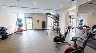 Located at 70 Ordnance St in Toronto's King West neighbourhood, this expansive fitness facility offers a well-appointed, contemporary exercise environment across a generous single floor. The space is thoughtfully designed with a modern aesthetic, featuring warm wood-panelled accent walls, recessed and cove lighting, and large floor-to-ceiling windows that flood the interior with natural light and provide views of the surrounding cityscape.

The main exercise area is anchored by a central artificial turf lane, ideal for functional training and sled work, complemented by rubber and carpeted flooring throughout the various zones. Cardio equipment — including treadmills, ellipticals, and rowing machines — lines the perimeter near the windows, while a dedicated strength zone houses a comprehensive array of resistance machines, cable systems, and free weights including dumbbells and medicine balls.

A separate, enclosed room provides space for cycling bikes, battle ropes, and additional free weights, offering a more intimate setting for group or focused training. Full-length mirrors are strategically placed to enhance the sense of space and support form-checking during workouts. The facility also benefits from direct access to an outdoor terrace, seamlessly connecting indoor training with the outdoors. The overall ambiance is clean, bright, and professional, reflecting a high standard of amenity design.