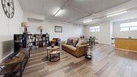 Located at 32 Ontario St in Kingston, Ontario, this social room serves as a comfortable communal gathering space spread across a single floor. The open-plan layout seamlessly connects a fully equipped kitchen with a relaxed living area, making it well-suited for group interaction and shared use.

The living area is furnished with a plush leather sofa, a dark leather armchair, and a pair of round wooden coffee tables arranged around a flat-screen television. A well-stocked bookcase adds a cozy, homey character to the space, while a large wall clock and decorative mirror contribute to its inviting ambiance. Track lighting mounted on the ceiling provides ample illumination throughout the room, complemented by natural light filtering in through windows along the walls.

The kitchen area features warm wood cabinetry, a granite-style countertop, and a central breakfast bar that doubles as a divider between the kitchen and living zones. Stainless steel appliances, including a full-size refrigerator and microwave, make this space practical for everyday use. The kitchen floor is finished in light ceramic tile, contrasting neatly with the wood-look laminate flooring that flows through the rest of the room.

Overall, the social room offers a functional, well-appointed environment ideal for residents to relax, socialize, and make use of shared facilities.