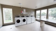 This shared laundry facility is a well-appointed, single-floor space designed with both functionality and comfort in mind. The room is finished with neutral taupe-toned walls and durable speckled vinyl flooring, creating a clean and practical environment. Multiple front-loading washing machines and top-load dryers are arranged along one wall, each equipped with individual coin-operated or card-reader controls, accompanied by posted usage instructions and informational notices for residents. A dedicated folding counter runs along the windowed wall, providing ample surface space for sorting and folding laundry. Large windows span much of one side of the room, allowing generous natural light to fill the space and offering views of surrounding greenery and the adjacent streetscape. Decorative framed artwork adds a welcoming touch to the walls, softening the utilitarian nature of the room. Accessed through a clearly labeled door, the laundry room connects to a carpeted corridor, making it easily accessible. The overall ambiance is bright, tidy, and thoughtfully organized, balancing practical amenities with an inviting atmosphere suited for everyday use.