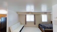 This single-floor space offers a versatile layout that combines living and sleeping areas within one open room. The interior is finished with neutral beige walls and smooth white ceilings, creating a clean and understated aesthetic throughout. A central window fitted with white blinds and complementary brown curtain panels allows natural light to filter into the room, contributing to a bright and welcoming atmosphere. A secondary window along the adjacent wall further enhances the natural illumination. The ceiling incorporates recessed HVAC vents, ensuring efficient climate control throughout the space. Flooring is covered with a light-toned area rug featuring a subtle geometric pattern, adding texture and visual interest underfoot. The room accommodates a white tufted sofa positioned along one wall, providing a seating area that complements the sleeping zone anchored by a full-sized bed with dark-toned bedding. The overall design is minimal and functional, with soft color choices and straightforward furnishings that contribute to a calm, practical living environment suited for everyday use.