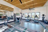 Located at 7631 Calloway Dr in Bakersfield, CA, this fitness center is a thoughtfully designed exercise space that combines modern aesthetics with a comprehensive range of workout equipment. The room's most distinctive architectural feature is its striking slatted wood ceiling, which adds warmth and a contemporary flair while housing recessed lighting and large ceiling fans that ensure comfortable air circulation throughout.

Tall, multi-pane windows line the walls, flooding the space with generous natural light and offering views of the surrounding neighborhood, including swaying palm trees. The bright, open atmosphere is further enhanced by crisp white walls and patterned modular carpet tiles in shades of teal, gray, and green, which define workout zones across the floor.

The exercise room is well-stocked with a variety of cardio equipment, including multiple treadmills, stationary spin bikes, an elliptical trainer, and a rowing machine. A dedicated free weights area features a full dumbbell rack alongside a weight bench, and TRX suspension training equipment is also available. Large wall-mounted mirrors expand the sense of space and allow users to monitor their form during workouts. The overall ambiance is energizing and modern, providing a fully functional fitness environment suited to a wide range of exercise routines.