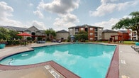 Located at 9955 Buffalo Speedway in Houston, Texas, this outdoor amenity space centers around a generously sized resort-style swimming pool with striking turquoise water and brick-trimmed edges. The pool area is designed for both relaxation and recreation, offering multiple entry points, including a built-in step entry and handrail access. Surrounding the pool is a spacious concrete sundeck furnished with lounge chairs and chaise seating, ideal for soaking up the Texas sun. Multiple shaded areas are provided through a combination of a covered gazebo and large market umbrellas in both blue and red, accommodating outdoor dining and casual gatherings at bistro-style table and chair sets. The landscaping throughout the space is well-maintained, featuring mature trees, trimmed hedges, colorful plantings, and decorative ceramic pots that add vibrant accents to the grounds. A brick-accented clubhouse with a metal roof and arched window serves as a architectural focal point at one end of the pool, adding a classic residential character to the setting. The surrounding multi-story residential buildings, constructed with a mix of brick and neutral siding, frame the space and provide a sense of enclosure and community. The overall ambiance is inviting and polished, offering a well-appointed outdoor retreat suited for everyday enjoyment.