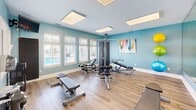 Located at 1314 Kingston Way in Chesapeake, Virginia, this fitness center offers a spacious, single-floor exercise room designed to support a comprehensive workout experience. The room is finished with light hardwood-style flooring and painted in a calming blue palette, creating an energizing yet welcoming atmosphere.

Large, multi-pane windows with white plantation shutters line multiple walls, flooding the space with abundant natural light and providing views of the surrounding outdoor area, including what appears to be a pool. Oversized flush ceiling light panels provide consistent, bright overhead illumination throughout.

A full-length mirrored wall expands the sense of space and supports proper form during exercise. The cardio zone is well-appointed with Matrix-brand treadmills, an elliptical, and a stationary bike. The strength training area features a multi-station cable machine, adjustable workout benches, and a rack of dumbbells. Stability and medicine balls in multiple sizes round out the functional fitness offerings.

A dedicated open area with open floor space provides room for stretching or floor-based exercises, while colorful artwork on the walls adds a touch of personality. A secondary adjoining area connects through a doorway, offering additional utility. This thoughtfully designed fitness center provides residents with a complete, accessible facility for their daily wellness routines.