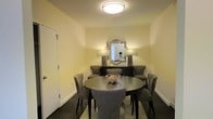 The dining area serves as a focal point of this single-story space, showcasing a circular dark-finished dining table surrounded by upholstered grey chairs with sleek, tapered legs. The room is finished with warm yellow-toned walls and dark hardwood flooring, creating a cozy yet sophisticated atmosphere. A flush-mounted circular ceiling light provides overhead illumination, complemented by two matching table lamps flanking an ornate, decoratively framed mirror mounted above a dark console table. The mirror's quatrefoil-inspired frame adds a touch of elegance to the space. A sculptural centerpiece rests atop the dining table, adding an artistic detail to the setting. The clean white baseboards and a white paneled door on the left wall contrast cleanly against the warm wall tones. The overall design reflects a cohesive, contemporary-classic aesthetic, with carefully selected furnishings that balance functionality and style. The intimate scale of the dining area is well-suited for gatherings, with thoughtful layering of light sources contributing to a warm and inviting ambiance.