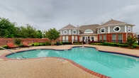 This residential community amenity area centers around a generously sized freeform swimming pool with a distinctive brick-trimmed coping and aggregate concrete surround. The pool's curvilinear design creates multiple zones for both active swimming and leisurely wading, with clearly marked depth indicators along the edges. Flanking the pool, the expansive deck offers ample space for outdoor furniture, including cushioned lounge chairs and bistro-style seating sets, ideal for relaxing poolside.

The community clubhouse serves as the architectural focal point, featuring a classic brick exterior, hipped rooflines, decorative circular windows, and arched dormers that lend a refined, traditional character to the space. Mature trees and well-maintained shrubs are thoughtfully arranged throughout the grounds, providing natural shade and greenery that soften the hardscape areas.

A dedicated children's playground is situated within the enclosed perimeter, offering colorful play structures complete with slides, making it a family-friendly addition to the amenity space. The entire area is enclosed by a wrought-iron fence, ensuring safety and privacy while maintaining open sightlines to the surrounding neighborhood. Decorative lamp posts are positioned throughout the grounds, providing ambient lighting for evening use. The overall atmosphere is welcoming, functional, and well-maintained.