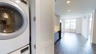 This single-floor unit offers a well-designed layout that maximizes functionality within a compact footprint. A louvered bifold door conceals a stacked washer and dryer unit, providing the convenience of in-unit laundry without sacrificing living space. The kitchen is outfitted with gray cabinetry, light countertops, stainless steel appliances including a French door refrigerator, and a dishwasher, all contributing to a clean, modern aesthetic. Dark hardwood-style flooring runs throughout, creating a warm contrast against the bright, neutral walls and recessed lighting overhead. The living area benefits from generous natural light courtesy of expansive floor-to-ceiling windows that frame views of the surrounding neighborhood and an adjoining outdoor space, such as a balcony or terrace. The open sightlines between the kitchen and living area enhance the sense of space, while the contemporary finishes and efficient layout make this unit both stylish and practical. Recessed ceiling lights provide consistent illumination throughout, complementing the abundant natural light from the large windows.