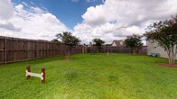 This community amenity space combines an outdoor dog park with a fully outfitted fitness center, providing residents with dedicated areas for both pet recreation and personal wellness. The dog park features a generously sized, fully fenced grassy area enclosed by tall wooden privacy fencing, offering a secure environment for pets to roam freely. Young trees are planted throughout the space, and a bench is available for owners. Playful bone-shaped signage marks the entrance, adding a welcoming touch. The surrounding neighborhood is visible beyond the fence line, giving the area an open, connected feel under an expansive sky. The fitness center is divided into distinct workout zones, all unified by stylish herringbone-patterned carpet flooring. The cardio area is bright and airy, featuring multiple treadmills positioned beside large windows with natural light pouring in, along with a wall-mounted television. The strength training zone is comprehensively equipped with a power rack, cable machine, rowing ergometer, and a Precor stationary bike. A dedicated functional training area houses a multi-purpose rig loaded with kettlebells, medicine balls, battle ropes, and resistance bands, complemented by a full-length mirror wall and ceiling fans that keep the space comfortable during intense workouts. A complete dumbbell rack rounds out the available equipment, making this a thorough and versatile training facility.
