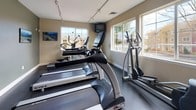 This fitness center is a dedicated, single-floor exercise facility designed to support a well-rounded cardio workout routine. The space features a dark, durable rubber flooring that is both practical and easy to maintain, complemented by neutral-toned walls that create a clean, focused atmosphere.

The main workout area is furnished with multiple True and Life Fitness brand treadmills arranged in a row, providing ample space for simultaneous use. An elliptical trainer and a stationary upright bike round out the cardio offerings, ensuring a variety of low-impact and high-intensity options for users of varying fitness levels. Wall-mounted televisions are positioned for easy viewing during workouts.

Natural light fills the space through several large windows that frame views of the surrounding outdoor environment, contributing to an open and energizing ambiance. Track lighting installed along the ceiling provides consistent, even illumination throughout the room. The entry features a classic white paneled door, and a small sanitation station with a dispenser and waste bin promotes cleanliness. Framed athletic artwork adorns the walls, reinforcing the fitness-oriented character of the space. Overall, the facility is thoughtfully arranged to maximize usability while maintaining a comfortable and motivating workout environment.