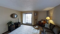 This single-floor space centers around a well-appointed living room designed for comfort and functionality. The room features wall-to-wall carpeting in a neutral tone, creating a soft and inviting foundation for the seating arrangement, which includes a plush sofa, an upholstered armchair with decorative pillows, and a tray-style coffee table.

A prominent sliding glass door serves as the focal point of the main wall, allowing natural light to fill the room while providing direct access to a private balcony with an outdoor view. Vertical blinds accompany the sliding door, offering adjustable light control. A wall-mounted air conditioning unit is positioned nearby, ensuring climate comfort year-round.

The walls are finished in a warm beige tone, complemented by a textured popcorn ceiling. Decorative touches include a round rope-framed mirror and a dark media console with decorative storage drawers along one wall. Baseboard heating is also visible, indicating a dual climate system. The overall ambiance is cozy and practical, with a layout that balances open living space with functional design elements.