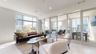 Located at 425 W Trade St in uptown Charlotte, NC, this contemporary amenity space offers a refined and functional environment designed for both relaxation and socializing. The interior is defined by clean lines, light-toned tile flooring, and crisp white walls that create a bright, airy atmosphere throughout.

The main lounge area is generously proportioned, anchored by a dark leather sofa, sculptural boucle accent chairs, and a solid wood coffee table. Large floor-to-ceiling windows flood the space with abundant natural light while framing panoramic views of the Charlotte skyline and surrounding greenery. Seamless glass partition doors open directly onto a spacious outdoor terrace, extending the living area and providing an al fresco setting furnished for lounging and dining.

A dedicated media and seating nook features a wall-mounted flat-screen display, a sleek console, and coordinated accent lighting, offering a more intimate gathering spot within the larger space.

The wet bar and dining area is outfitted with sophisticated grey cabinetry, a textured tile backsplash, natural wood wall paneling, a built-in beverage refrigerator, and ample counter space — ideal for entertaining. A large dining table with upholstered chairs completes the zone. Together, these areas deliver a polished, urban retreat in the heart of Charlotte.