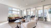 Located at 425 W Trade St in uptown Charlotte, NC, this contemporary amenity space offers a refined and functional environment designed for both relaxation and socializing. The interior is defined by clean lines, light-toned tile flooring, and crisp white walls that create a bright, airy atmosphere throughout.

The main lounge area is generously proportioned, anchored by a dark leather sofa, sculptural boucle accent chairs, and a solid wood coffee table. Large floor-to-ceiling windows flood the space with abundant natural light while framing panoramic views of the Charlotte skyline and surrounding greenery. Seamless glass partition doors open directly onto a spacious outdoor terrace, extending the living area and providing an al fresco setting furnished for lounging and dining.

A dedicated media and seating nook features a wall-mounted flat-screen display, a sleek console, and coordinated accent lighting, offering a more intimate gathering spot within the larger space.

The wet bar and dining area is outfitted with sophisticated grey cabinetry, a textured tile backsplash, natural wood wall paneling, a built-in beverage refrigerator, and ample counter space — ideal for entertaining. A large dining table with upholstered chairs completes the zone. Together, these areas deliver a polished, urban retreat in the heart of Charlotte.