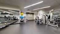 Located at 7 DeKalb Avenue in Brooklyn, New York, this fitness facility offers a comprehensive, single-floor gym experience designed to accommodate a wide range of workout styles and fitness goals. The space is thoughtfully organized into distinct training zones, each outfitted with professional-grade equipment.

The cardio area features a generous row of treadmills and a stair climber, all set against full-length mirrors that amplify the sense of space. Linear LED lighting runs across the drop-tile ceiling, providing bright, even illumination throughout. A dedicated cardio alcove houses elliptical trainers and upright cycling bikes, with wall-mounted televisions for an engaging workout experience.

The strength and functional training zone includes a cable machine, plate-loaded strength equipment, barbells, weight plates, and foam rollers, all set on durable rubber flooring. A rack of stability balls in multiple sizes adds versatility for core and balance training. Mirrored walls throughout these areas allow members to monitor their form.

A separate multi-purpose studio with glass partition walls and doors offers a clean, open floor plan ideal for stretching, group classes, or rowing machine workouts. A ballet barre along one mirrored wall further extends the studio's functionality. The overall design is clean and modern, with white walls, neutral tones, and a well-maintained interior that creates a focused and professional training environment.