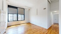 This single-floor residence offers four bedrooms, two bathrooms, a living room, and a kitchen, all connected by two hallways and unified by warm hardwood flooring throughout. The layout makes efficient use of its footprint, with the spacious primary bedroom offering generous room for furnishings.

Large, black-framed windows are a defining architectural feature across multiple rooms, flooding the interiors with natural light and framing urban cityscape views. Exposed brick detailing visible at window surrounds adds an industrial character that complements the clean white walls and textured ceilings.

The kitchen stands out with dark cabinetry, white quartz countertops, a subway tile backsplash, open wood shelving, pendant lighting, and a full suite of stainless steel appliances including a gas range, microwave, dishwasher, and refrigerator. The bathrooms feature white large-format subway tile with glass-panel shower enclosures and matte black hardware, reflecting a modern aesthetic. The secondary bedrooms are compact but well-lit, each benefiting from the abundant natural light provided by the oversized windows. The overall ambiance is clean, modern, and urban.