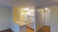 This single-story residence includes a kitchen designed for efficient use of space. The kitchen is defined by white cabinetry that lines the upper walls, providing ample storage, while a set of white appliances — including a refrigerator, electric range with coil burners, and a single-basin sink — are neatly arranged along the back wall. A flush-mounted fluorescent light fixture illuminates the workspace, highlighting the light-colored walls and wood-look flooring that runs through the kitchen area. A peninsula-style breakfast bar extends from one side, creating a natural divide between the kitchen and the adjacent living space, which features carpeted flooring. The textured ceiling adds a classic residential touch throughout. A hallway visible from the kitchen leads further into the home, suggesting additional rooms beyond. The overall layout is practical and straightforward, offering a cohesive, easy-to-navigate floor plan suited for everyday living.