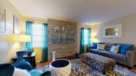 This single-story home presents a cohesive and thoughtfully designed interior highlighted by a bold reclaimed wood accent wall that serves as the living room's focal point. The living area is generously sized, featuring warm hardwood flooring, large windows dressed in vibrant teal curtains that invite natural light, and a brick-trimmed base beneath the accent wall that adds architectural character. A tufted ottoman and coordinated furnishings contribute to the room's inviting ambiance.

Adjacent to the living area, the dining space connects seamlessly to the kitchen, which features white cabinetry, dark countertops, and hardwood flooring that continues throughout. A pendant light over the dining table adds a refined touch to this open, functional area.

The primary bedroom offers plush carpeting, a ceiling fan, and direct access to a private en-suite bathroom with a vanity and walk-in closet. A secondary bedroom provides comfortable accommodations with carpeted flooring and a bright window. Both bathrooms are finished with dark granite-style countertops and tub/shower combinations.

A dedicated laundry area houses a full-sized washer and dryer, adding everyday convenience. The consistent teal and neutral color palette throughout creates a unified, polished atmosphere across all spaces.
