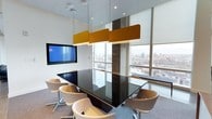 This professional meeting room offers a refined and functional environment ideal for focused collaboration and presentations. The space is anchored by a large, glossy dark conference table surrounded by contemporary cream-upholstered swivel chairs, providing comfortable seating for multiple attendees. Striking gold acoustic pendant panels hang overhead, adding both a bold design element and sound management functionality to the room. Recessed lighting complements the natural light that floods in through expansive floor-to-ceiling windows lining the walls, offering sweeping panoramic views of the city skyline beyond. A wall-mounted flat-screen display is prominently positioned for presentations and video conferencing, ensuring the room is fully equipped for modern business needs. The neutral color palette — featuring white walls, grey carpet, and clean architectural finishes — creates a calm, professional ambiance that keeps the focus on the work at hand. The combination of natural light, city views, thoughtful acoustics, and high-quality furnishings makes this meeting room a sophisticated and practical space for professional gatherings of all kinds.