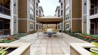 This outdoor communal space is thoughtfully designed to offer residents a welcoming and functional environment centered around a spacious, large-format tiled courtyard. At its heart, a freestanding canopy shelters a dining table and chairs, providing an ideal setting for outdoor gatherings, while a nearby grill station adds convenient amenity for al fresco cooking and entertaining. Raised stone planters line the walkways, filled with tropical palms and lush, variegated foliage that introduce vibrant greenery throughout the space. The courtyard is flanked on both sides by multi-story residential buildings clad in warm taupe horizontal siding with brick accents, creating an enclosed yet open-air atmosphere. A covered entrance corridor features exposed dark wood beam detailing overhead, recessed lighting, and decorative metal railing with a cross-hatch pattern, lending an architectural elegance to the transitional areas. From this covered walkway, views open to a sparkling pool area below, enhancing the resort-like character of the space. The integration of mature landscaping, stone-trimmed planters, and wide tiled pathways ensures both visual appeal and ease of movement throughout, making this courtyard a defining communal feature of the property.