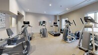 This fitness facility is organized across a single level, comprising two interconnected workout areas designed to support a full range of exercise routines. The space features a practical and functional interior with drop-panel ceilings, durable rubber flooring in the strength training zone, and carpeted flooring in the cardio area, providing appropriate surfaces for each type of activity.

The strength training room is outfitted with a power rack, adjustable weight bench, Olympic plates, dumbbells on a dedicated rack, and a cable machine, making it suitable for a wide variety of resistance exercises. Large wall mirrors enhance the sense of space and allow users to monitor their form. Wood-panel wainscoting along the lower walls adds a structured visual element throughout. A wall-mounted flat-screen TV and a first aid station contribute to both comfort and safety.

The cardio zone is a bright, well-lit room featuring recessed lighting and a window that introduces natural light. It houses multiple treadmills, elliptical trainers, stair climbers, and stationary bikes, along with a wall-mounted TV and exercise reference posters. The overall ambiance is clean, practical, and purposefully designed to accommodate a diverse set of fitness needs in a compact yet efficiently utilized space.