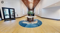 Located at 330 Towerview Ct in Cary, NC, this expansive single-floor amenity space offers a refined and contemporary environment across a generous footprint. The grand entrance hallway immediately sets the tone, featuring soaring double-height ceilings clad in warm wood paneling, light hardwood flooring, and a dramatic sculptural light fixture. A central marble-topped table with fluted wood legs sits atop a bold circular blue area rug, creating a striking focal point upon entry. Floor-to-ceiling black-framed windows flood the space with natural light and offer views of the surrounding landscaped courtyard. The open living area is elegantly appointed with neutral-toned furnishings, large-scale marble accent walls with gold veining, and tray ceilings with recessed wood detailing. The dining area features a curved island bar with counter seating, pendant lighting, and vertical wood slat wall accents. A dedicated workspace zone includes a long window-side desk with live greenery, complemented by bold teal built-in shelving. A private conference room offers an intimate setting with frosted glass partition walls, a central meeting table, leather chairs, and cascading gold pendant lights. Living moss wall art installations add organic texture throughout, completing this thoughtfully designed, upscale communal environment.