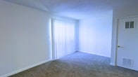 This single-floor space offers a clean, functional layout with a straightforward design that prioritizes comfort and practicality. The interior features wall-to-wall carpeting in a neutral tone, providing a soft and quiet underfoot experience throughout. Crisp white walls create a bright, open atmosphere, while white baseboard trim adds a clean finishing touch to the room's perimeter.

A prominent floor-to-ceiling window fitted with vertical blinds serves as the room's focal point, allowing an abundance of natural light to flood the space. When the blinds are drawn, they diffuse incoming sunlight into a soft, even glow that illuminates the entire room. The window's large scale enhances the sense of openness and airiness within the space.

The ceiling features a textured finish, adding subtle visual depth overhead. The overall ambiance is calm and neutral, offering a versatile blank canvas well-suited to a variety of uses and furnishing styles. The minimalist design ensures that the space feels uncluttered and easy to personalize.