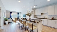 This single-floor residence is designed with a contemporary open-concept layout that seamlessly connects the kitchen, dining, and living areas. The kitchen is a standout feature, outfitted with sleek white cabinetry, dark countertops, a classic subway tile backsplash, and a full suite of stainless steel appliances. A generously sized island with a dark wood-toned countertop provides seating for multiple guests and serves as a natural gathering point. Pendant lighting above the island adds both function and visual interest.

The living area is warm and inviting, featuring light hardwood-style flooring, neutral taupe walls, and large windows dressed with navy drapes that allow abundant natural light to fill the space. The layout flows naturally from the kitchen into this relaxed seating area.

The primary bedroom features large windows with outdoor views, soft carpeting, and a calming neutral palette. A second bedroom showcases a bold blue accent wall paired with ample natural light. The bathroom is clean and functional, finished with white cabinetry, dark countertops, and hardwood-style flooring consistent with the rest of the residence. Throughout, the design balances modern finishes with a warm, livable aesthetic.