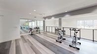 Located in Frisco, TX, this well-appointed two-story fitness center offers a comprehensive workout environment designed to support a wide range of exercise disciplines. The lower level features patterned carpet tiles in grey and yellow-green tones, lending the space a contemporary aesthetic. This floor houses a functional training station with cable machine attachments, plyometric boxes, open shelving for accessories, and additional strength equipment. Decorative wall art adds a refined touch to the training environment. A staircase connects to the upper level, which transitions to sleek striped luxury vinyl plank flooring. This floor is dedicated to cardio and group fitness, featuring a row of spinning bikes, kettlebell racks, stability balls, and medicine balls neatly organized along the walls. Floor-to-ceiling windows along the perimeter of the facility flood both levels with natural light and offer views of the outdoor surroundings, including a pool area. Large mirrors extend the visual depth of the cardio zone, which is lined with Matrix-brand treadmills, ellipticals, and stair climbers, each equipped with individual monitors. Recessed lighting and high ceilings contribute to the open, airy atmosphere throughout the facility.