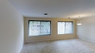 This single-floor space presents a clean, straightforward layout centered around a spacious main room that is both functional and welcoming. The room is finished with wall-to-wall neutral-toned carpeting, providing a soft underfoot feel that contributes to a calm and comfortable atmosphere. The walls are painted in a light, warm beige tone, enhancing the sense of openness throughout.

A defining feature of the main room is its generous window arrangement. Two large picture windows, each fitted with horizontal mini-blinds for adjustable light control, line the exterior wall and allow abundant natural light to fill the interior. These windows frame views of mature trees and a neighboring multi-story building, giving the space a pleasant connection to the outdoors. A third, slightly smaller window further contributes to the bright and airy character of the room.

The ceiling is smooth and white, fitted with a centrally placed ventilation grille, maintaining a tidy and uncluttered overhead appearance. The clean white baseboard trim provides a crisp contrast against the carpeted floor, rounding out the simple yet polished interior finishes. Overall, this space offers a neutral, light-filled environment well-suited for a variety of uses.