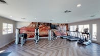 This well-appointed fitness center offers a thoughtfully designed workout environment that combines functionality with striking visual appeal. The space is anchored by a dramatic, large-scale photographic mural depicting a rugged desert canyon landscape in vivid red and orange tones, creating an energizing focal point along one wall. A mounted flat-screen television is integrated into the mural wall, adding entertainment options during workouts.

The flooring features a sophisticated herringbone-patterned wood-look surface in a neutral gray tone, lending the space a contemporary, polished aesthetic. Recessed lighting is evenly distributed across the white ceiling, providing consistent, well-lit conditions throughout the room.

A generous lineup of cardio equipment includes multiple treadmills and an elliptical trainer, while strength training needs are met with a multi-station cable machine and a dedicated weight bench. The equipment is arranged to allow ample circulation space between stations.

Along the far wall, a row of full-height glass doors and large windows floods the space with natural light and provides views of the outdoor area beyond, creating an open, airy atmosphere that enhances the overall workout experience.