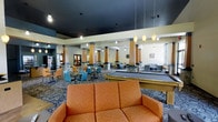 This single-floor community gathering space is designed to accommodate a variety of social and recreational activities. The expansive open layout is anchored by a series of decorative columns with warm wood-toned accents, lending a sense of structure and architectural character to the room. A dark, dramatic ceiling contrasts with the lighter walls, creating a dynamic visual atmosphere throughout the space.

At the center of the room sits a full-sized billiards table, making it a focal point for recreation. Surrounding it are multiple casual seating arrangements, including comfortable upholstered sofas in warm tones, as well as numerous dining-height tables paired with bright teal chairs, offering ample space for group gatherings or individual relaxation.

Pendant lighting hangs from the ceiling in clusters, providing warm, ambient illumination that complements the decorative patterned carpet underfoot. Along the perimeter, additional seating areas offer quieter spaces for conversation or leisure. A display or bookshelf area is visible near the entrance, suggesting resources for residents or visitors. Large doors and windows allow natural light to filter into certain sections, enhancing the overall openness of the space. The combination of recreational amenities, varied seating zones, and thoughtful design details makes this a well-appointed and versatile community space.