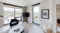 This single-floor unit offers a thoughtfully designed open-concept layout with a clean, contemporary aesthetic. Light wood flooring runs throughout the main living areas, complementing the neutral white walls and creating a bright, cohesive atmosphere.

The kitchen is fitted with flat-front light wood cabinetry, dark countertops, and integrated appliances including an oven and cooktop. A long wood dining table with bench seating and modern pendant lighting connects the kitchen to the living area, maximizing the functional use of space.

The living room is a standout area, featuring multiple floor-to-ceiling windows that frame sweeping panoramic views of the cityscape and surrounding hills. The space accommodates a plush sofa, leather armchair, and a mounted television positioned on a sculptural concrete column.

The bedroom offers a serene retreat with soft carpeting, a woven upholstered headboard, dual wall-mounted sconces, and a large window providing continued views of the hillside. A walk-in closet is accessible from the bedroom.

The bathroom features stone tile flooring, a glass-enclosed walk-in shower with a rainfall showerhead, an open-shelf vanity, and an organically shaped mirror with flanking wall sconces, delivering a refined, spa-inspired finish.