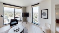 This single-floor unit offers a thoughtfully designed open-concept layout with a clean, contemporary aesthetic. Light wood flooring runs throughout the main living areas, complementing the neutral white walls and creating a bright, cohesive atmosphere.

The kitchen is fitted with flat-front light wood cabinetry, dark countertops, and integrated appliances including an oven and cooktop. A long wood dining table with bench seating and modern pendant lighting connects the kitchen to the living area, maximizing the functional use of space.

The living room is a standout area, featuring multiple floor-to-ceiling windows that frame sweeping panoramic views of the cityscape and surrounding hills. The space accommodates a plush sofa, leather armchair, and a mounted television positioned on a sculptural concrete column.

The bedroom offers a serene retreat with soft carpeting, a woven upholstered headboard, dual wall-mounted sconces, and a large window providing continued views of the hillside. A walk-in closet is accessible from the bedroom.

The bathroom features stone tile flooring, a glass-enclosed walk-in shower with a rainfall showerhead, an open-shelf vanity, and an organically shaped mirror with flanking wall sconces, delivering a refined, spa-inspired finish.