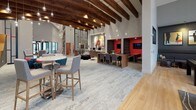 This single-story clubhouse is a thoughtfully designed communal space that blends modern aesthetics with versatile functionality. The architecture is defined by dramatic vaulted ceilings adorned with rich, dark-stained exposed wood beams, from which pendant globe lights hang at varying heights, creating a warm and inviting ambiance across the open floor plan. Large black-framed windows flood the interior with abundant natural light and provide views to the surrounding exterior.

The main lounge area features stylish patterned armchairs with bold accent pillows arranged around area rugs, complemented by high-top bistro tables for casual seating. A wall-mounted flat-screen display serves as a focal point in this zone. Adjacent to the lounge is a dedicated co-working area with built-in desks, desktop computers, sculptural egg-style chairs, and intimate red-accented booth alcoves equipped with screens for private meetings or focused work.

A café-style bar counter with a white quartz surface and upholstered bar stools faces a built-in credenza featuring dual monitors, a coffee station, and an intricate botanical wall-art backsplash. The game room offers a full-size billiards table, a tufted banquette beneath a gallery wall of contemporary art, and high-top seating beside floor-to-ceiling windows. Light wood-look flooring unifies the space, while curated artwork and bold color accents throughout contribute to an energetic yet refined atmosphere.