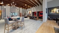 This single-story clubhouse is a thoughtfully designed communal space that blends modern aesthetics with versatile functionality. The architecture is defined by dramatic vaulted ceilings adorned with rich, dark-stained exposed wood beams, from which pendant globe lights hang at varying heights, creating a warm and inviting ambiance across the open floor plan. Large black-framed windows flood the interior with abundant natural light and provide views to the surrounding exterior.

The main lounge area features stylish patterned armchairs with bold accent pillows arranged around area rugs, complemented by high-top bistro tables for casual seating. A wall-mounted flat-screen display serves as a focal point in this zone. Adjacent to the lounge is a dedicated co-working area with built-in desks, desktop computers, sculptural egg-style chairs, and intimate red-accented booth alcoves equipped with screens for private meetings or focused work.

A café-style bar counter with a white quartz surface and upholstered bar stools faces a built-in credenza featuring dual monitors, a coffee station, and an intricate botanical wall-art backsplash. The game room offers a full-size billiards table, a tufted banquette beneath a gallery wall of contemporary art, and high-top seating beside floor-to-ceiling windows. Light wood-look flooring unifies the space, while curated artwork and bold color accents throughout contribute to an energetic yet refined atmosphere.