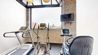 Located at 79 Warren St in Glens Falls, NY, this fitness center offers a well-appointed exercise space designed to support an active lifestyle. The room's most striking architectural feature is its exposed brick wall, which lends a warm, industrial character to the space while complementing the clean, white-painted walls on the opposing sides. Large windows along the brick wall flood the room with natural light and frame views of the surrounding Glens Falls neighborhood, creating an open and energizing atmosphere despite the room's intimate scale. A skylight-style ceiling structure further enhances the brightness of the space. The flooring consists of durable rubber gym flooring in a dark speckled finish, well-suited for high-traffic exercise use. The room is equipped with a treadmill, an elliptical trainer, and a recumbent stationary bike, providing a solid range of cardio workout options. A wall-mounted flat-screen television is positioned for easy viewing during workouts. A wall-mounted mini-split air conditioning unit ensures climate comfort year-round. The room is accessed through a dedicated entry door and is clearly identified as a fitness center. Overall, the space combines functional gym amenities with distinctive architectural charm.
