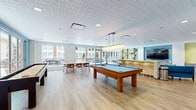 This single-floor amenity space is thoughtfully designed to serve as a dynamic social and recreational hub. The open layout centers around a full-size pool table beneath a statement pendant light, complemented by a shuffleboard table for additional entertainment. Light hardwood flooring flows throughout, unifying the various activity zones seamlessly.

A generously sized curved teal sofa anchors the lounge area, positioned beside a mosaic-tiled bar counter and a wall-mounted flat-screen TV. Built-in shelving with a bold yellow-green finish adds a playful pop of color and display space nearby. Large floor-to-ceiling windows and glass doors flood the interior with natural light while offering direct access to an outdoor terrace, providing views of the surrounding urban streetscape.

A dedicated dining area features a long communal table with mid-century-style chairs, positioned adjacent to additional windows overlooking the terrace. The kitchenette is well-equipped with stainless steel appliances, taupe flat-front cabinetry with gold hardware, and a vivid turquoise tile backsplash that adds a distinctive design accent.

A corridor with bold plaid wallcovering and a sculptural ceiling fixture leads to restrooms and a glass-fronted fitness center, rounding out the amenities of this energetic, well-appointed space.