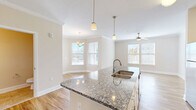 Located at 4005 Galantis Dr in Morehead City, NC, this well-appointed single-floor residence offers a thoughtfully designed layout with one bedroom, two bathrooms, and a range of functional spaces including a pantry, dedicated laundry room, utility room, hallway, and two closets. The heart of the home is an open-concept living, dining, and kitchen area bathed in natural light from multiple windows. The kitchen features a large granite-topped island with a double basin sink, white shaker cabinetry, stainless steel appliances including a refrigerator, range, and microwave, and elegant pendant lighting overhead. Crown molding adds a refined touch throughout the main living areas. The dining area is anchored by a stylish chandelier and benefits from a glass-panel exterior door with transom window, allowing abundant natural light. The spacious bedroom is finished with plush carpet, a ceiling fan, and a window with exterior views. Additional rooms display warm hardwood-style flooring and neutral toned walls throughout. Both bathrooms feature granite vanity tops and tile flooring. The dedicated laundry room comes equipped with a washer and dryer. This residence combines practical amenities with clean, contemporary finishes in a comfortable coastal Carolina setting.