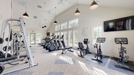 Located at 267 Grand Cypress Road in Summerville, SC, this fitness center is a standout amenity designed to support a full range of workout routines. The space is defined by its dramatic vaulted ceilings, which lend an open, airy atmosphere throughout. Large multi-pane windows line the walls, flooding the interior with natural light and offering views of the surrounding outdoor area, including a glimpse of a pool and lush greenery beyond the French doors.

The layout is thoughtfully organized into distinct zones. The cardio area features a row of elliptical machines, treadmills, a stationary upright bike, and a spin bike with an interactive display, all positioned to take advantage of the natural light from the expansive windows. The strength-training section is equipped with a multi-station cable machine, weight benches, and various resistance machines, complemented by floor-to-ceiling mirrors that enhance the sense of space.

Stylish pendant lighting with matte black fixtures and ceiling fans add to the clean, modern aesthetic. The flooring combines patterned carpet tiles with rubberized mats in the equipment zones. Motivational quotes are displayed on the walls, contributing to an energizing atmosphere. A dedicated entry area with French doors opens directly to the outdoor pool deck, seamlessly connecting indoor fitness with outdoor amenities.