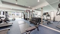 This single-level fitness facility offers a comprehensive and modern workout environment designed to support a wide range of exercise routines. The space seamlessly integrates both indoor and outdoor areas, creating a dynamic and inviting atmosphere for users.

The indoor fitness area is outfitted with blue carpeted flooring and features recessed lighting throughout, contributing to a clean and contemporary aesthetic. A dedicated cardio zone houses multiple treadmills and stationary bikes, each equipped with individual screens for an engaging workout experience. A free weights section includes a full dumbbell rack paired with an adjustable bench, while a cable machine system — including Precor equipment — provides versatile strength training options. Floor mats offer space for stretching and bodyweight exercises, and mirrored walls enhance the sense of openness throughout.

Large glass sliding doors and floor-to-ceiling windows allow natural light to flow into the fitness area and provide a visual connection to the outdoors. The exterior features synthetic turf alongside light-toned paver tiles, with raised planter walls adding a landscaped element to the space.

An adjacent glass-walled lounge area with wood-look flooring is visible from within the gym, offering a complementary space for relaxation or informal gatherings.