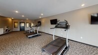 Located at 540 Tyler Rd S in Red Wing, Minnesota, this fitness center offers a generously proportioned, single-floor space designed to support a well-rounded exercise routine. The room is finished with durable patterned carpeting and neutral-toned walls accented by darker trim, creating a clean and functional aesthetic throughout.

Recessed lighting illuminates the space evenly, supplemented by multiple windows along the perimeter that allow natural light to filter in and provide views of the surrounding exterior. A wall-mounted flat-screen television is positioned for easy viewing from the cardio equipment area, enhancing the workout experience.

The facility is outfitted with a variety of cardio machines, including treadmills equipped with individual console screens, elliptical trainers, a recumbent stationary bike, and additional step and rowing equipment. The layout provides ample floor space between machines, allowing for comfortable movement throughout the room.

A built-in dual water fountain station is conveniently located near the exit door, offering easy hydration access. Framed wall panels near the entrance area add a structured, finished look. The space is accessed through a keyed door with clearly marked emergency exit signage, reflecting attention to safety and accessibility standards.