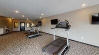 Located at 540 Tyler Rd S in Red Wing, Minnesota, this fitness center offers a generously proportioned, single-floor space designed to support a well-rounded exercise routine. The room is finished with durable patterned carpeting and neutral-toned walls accented by darker trim, creating a clean and functional aesthetic throughout.

Recessed lighting illuminates the space evenly, supplemented by multiple windows along the perimeter that allow natural light to filter in and provide views of the surrounding exterior. A wall-mounted flat-screen television is positioned for easy viewing from the cardio equipment area, enhancing the workout experience.

The facility is outfitted with a variety of cardio machines, including treadmills equipped with individual console screens, elliptical trainers, a recumbent stationary bike, and additional step and rowing equipment. The layout provides ample floor space between machines, allowing for comfortable movement throughout the room.

A built-in dual water fountain station is conveniently located near the exit door, offering easy hydration access. Framed wall panels near the entrance area add a structured, finished look. The space is accessed through a keyed door with clearly marked emergency exit signage, reflecting attention to safety and accessibility standards.