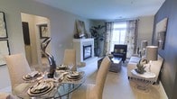This single-floor residence showcases a thoughtfully designed open-concept layout that seamlessly connects the dining and living areas, creating a cohesive and welcoming atmosphere. The living room centers around a classic white fireplace with a sleek surround, serving as a natural focal point and adding warmth to the space. Large windows dressed with patterned floral curtains allow generous natural light to brighten the room, enhancing the neutral, warm-toned color palette found throughout. Plush carpeting underfoot contributes to a comfortable and refined ambiance. The living area accommodates a well-arranged seating arrangement, including a sofa and accent chair positioned around a rectangular coffee table. Adjacent to the living room, the dining area features a round glass-top table set for entertaining, complemented by upholstered chairs and tasteful decorative accents, including a sculptural centerpiece. An arched passageway visible in the background hints at additional rooms, while tall indoor plants add organic texture. The overall design balances elegance with comfort, presenting a polished and livable environment.